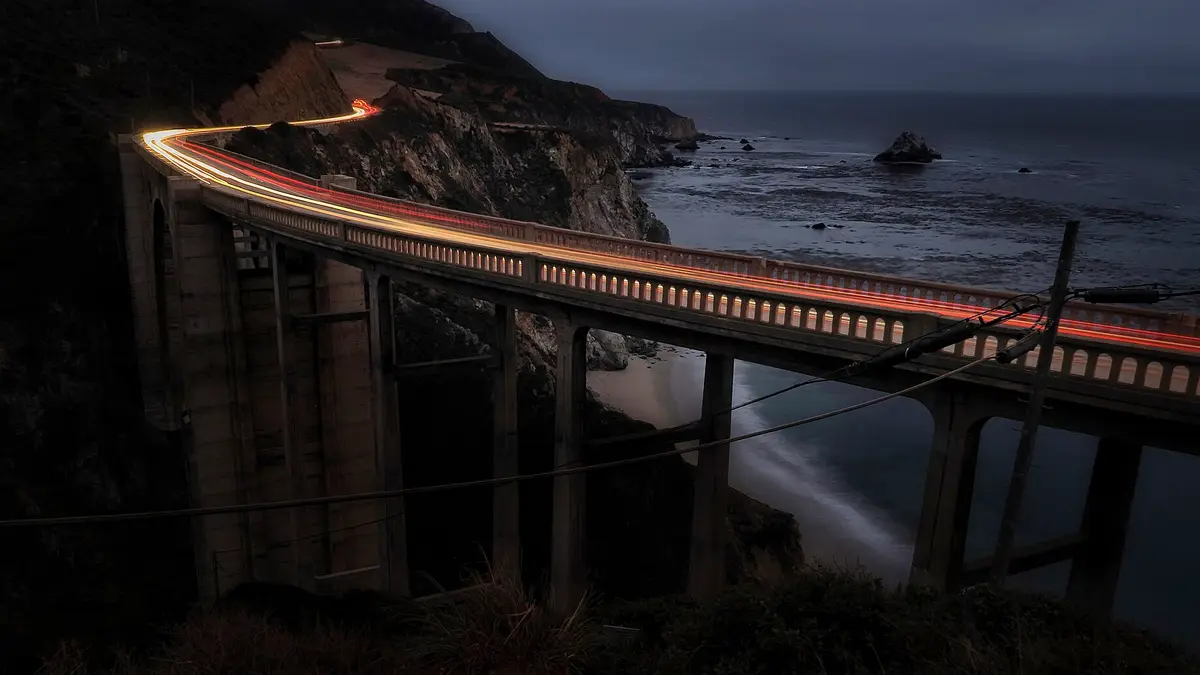 Puente Bixby en la costa de California de noche, con estelas de luz de los autos y el océano Pacífico al fondo