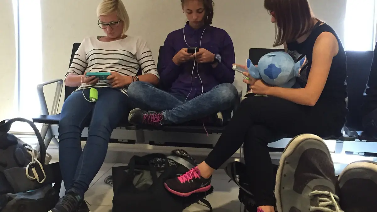 Tres jóvenes sentadas en una sala de embarque usando sus celulares