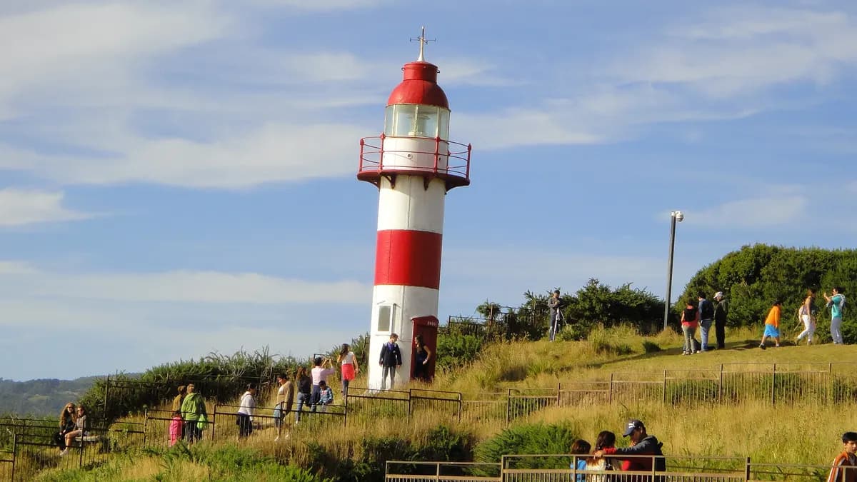 Visitantes junto al faro rojo y blanco de Niebla en una loma verde frente al mar