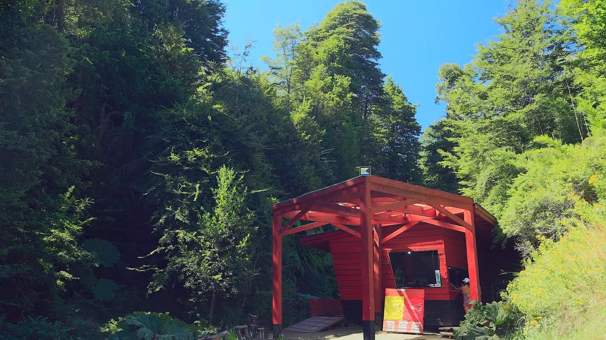 Pequeña construcción roja de ingreso a las termas entre árboles nativos